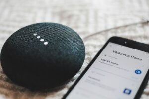 Google Home Mini smart speaker next to a smartphone, showing speakers still use Google Assistant for now