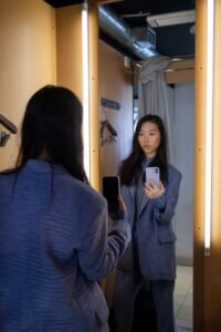 Woman taking a mirror selfie while trying on clothes in a fitting room.